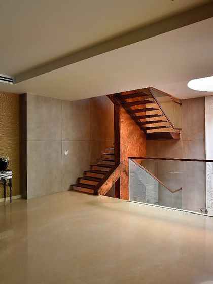This view showcases the main landing of a Delhi farmhouse, where modern and spiritual elements converge. A floating wooden staircase is juxtaposed with a serene white bas-relief wall panel depicting lotuses, all illuminated by a circular skylight that bathes the space in soft, natural light.
