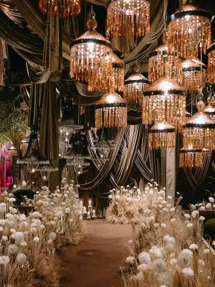 An ethereal Sangeet decor theme, with a pathway lined with dried grasses and white pom-pom flowers, under a canopy of crystal chandeliers. This design blends rustic and glamorous elements for a unique look.