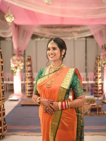 A full-length shot of the bride at her wedding venue. The traditional orange saree with a green border stands out beautifully.