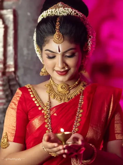 A gentle and serene portrait. The bride holds a small diya, her gaze directed downwards, creating a moment of quiet prayer and contemplation. The pink backlighting adds a soft, ethereal glow.