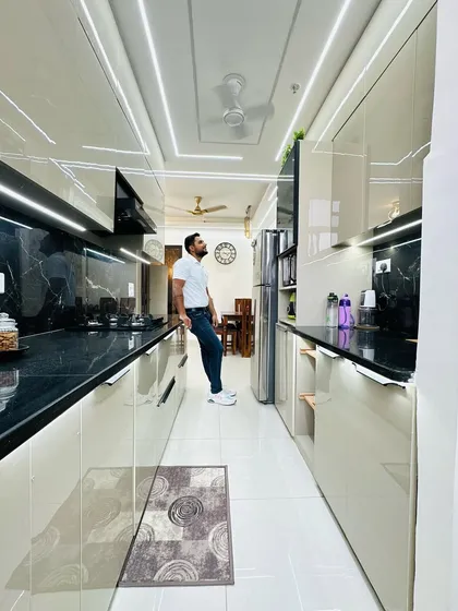 This is a parallel modular kitchen with a balcony extension, designed for a client at Nirala Estate. The kitchen features beige high-gloss acrylic cabinets, a black marble countertop, and profile lighting for a bright and functional cooking space.