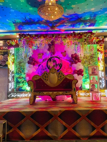 A royal stage setup for a Naming Ceremony in a banquet hall. The decoration features a throne-like sofa for the baby, balloon pillars, and a personalized photo display.