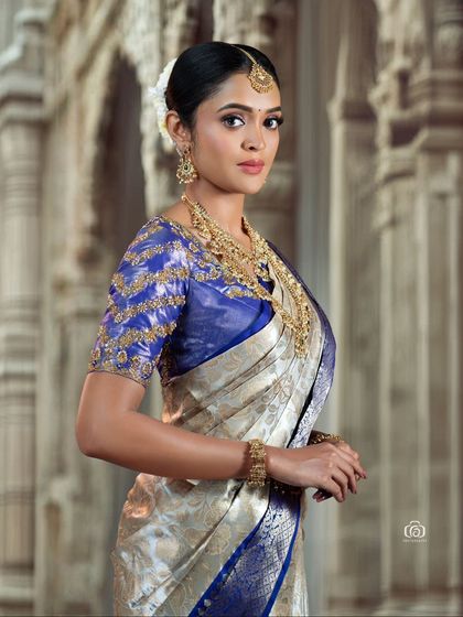 A stunning bridal portrait featuring a silver and blue Kanjivaram saree. The background, reminiscent of temple architecture, adds a sense of grandeur to this traditional South Indian look.