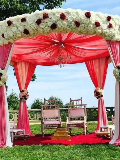 An elegant outdoor Vidhi Mandap we designed at a resort, featuring a beautiful floral canopy of white and red roses with flowing pink drapes. We create sacred and beautiful spaces for you to take your vows.