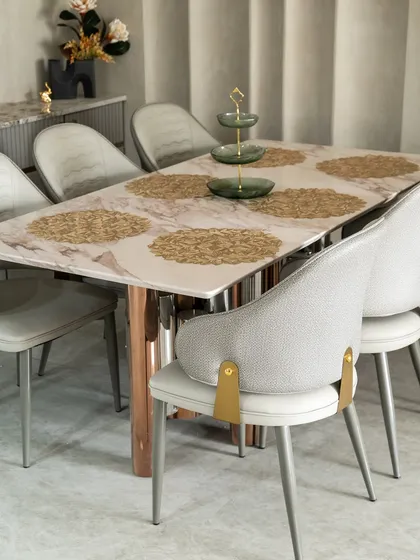 This dining setup exudes quiet luxury. The marble tabletop with soft pink veining is paired with chairs accented by brushed gold brackets. Every detail, from the golden placemats to the tiered centerpiece, is curated for a refined dining experience.