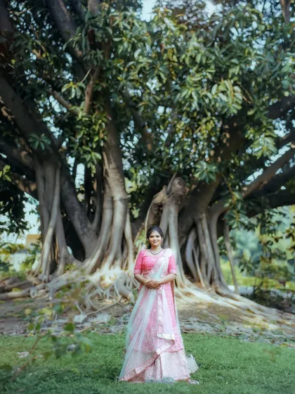 A full-length portrait showing the scale of the massive banyan tree. The woman in her beautiful pink lehenga stands in contrast to the ancient, powerful nature surrounding her.