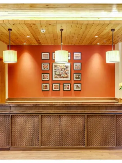The reception desk at Janjehli Resort features intricate wooden carving and is set against a bold red wall adorned with traditional Chamba Rumal artwork. This immediately immerses guests in the local culture.