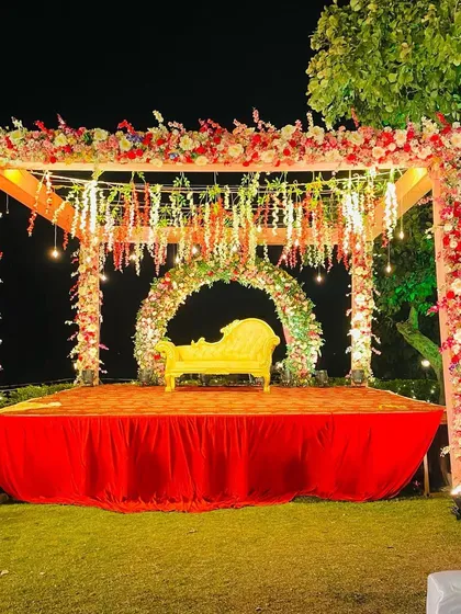 An outdoor ring ceremony stage designed to feel both grand and natural. The mandap is decorated with garlands of fresh flowers, hanging lights, and a floral arch backdrop, all set against the beautiful evening sky.