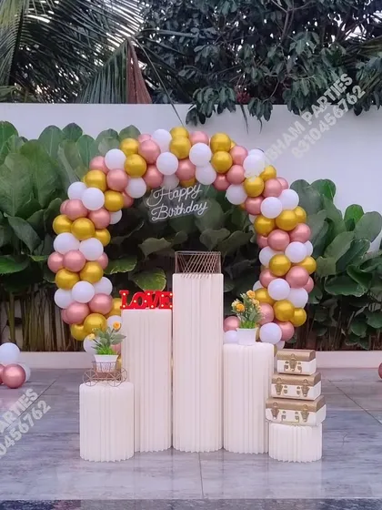 An elegant outdoor birthday setup featuring a circular balloon arch in rose gold, white, and gold, with stylish plinths for the cake.