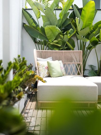 This sun-drenched balcony was transformed with large tropical leaves for privacy and visual height. The combination of wooden decking, soft seating, and curated greenery makes it a true outdoor room.