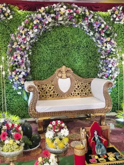 A baby shower stage featuring a mix of real and artificial elements. The green foliage wall is brightened by a colorful floral arch, with some of the Seer Thattu gift baskets visible in the front.