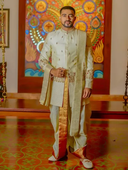 This full-length view showcases a complete South Indian groom's outfit. The off-white layered sherwani features subtle embroidery and is paired with a traditional dhoti with a gold border, creating a look of pure elegance.