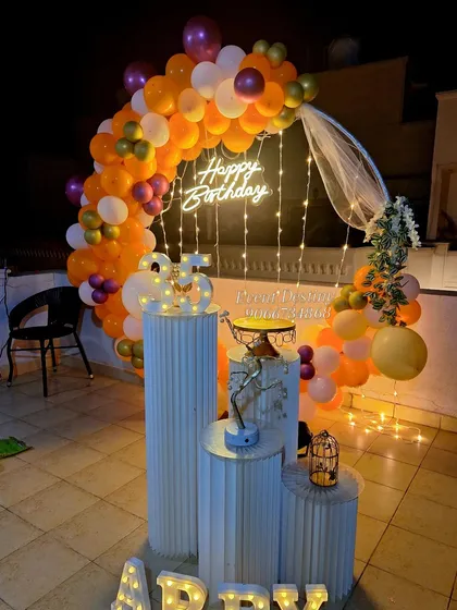 A vibrant rooftop birthday surprise with a circular balloon arch in orange and purple. The setup includes a custom 'Happy Birthday' neon sign and tiered pedestals for the cake and decor.