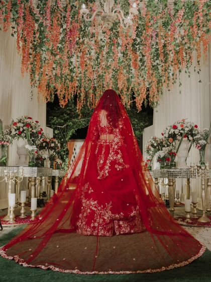 A breathtaking shot of the bride from behind, showcasing her long, magnificent red veil against a beautifully decorated floral mandap.