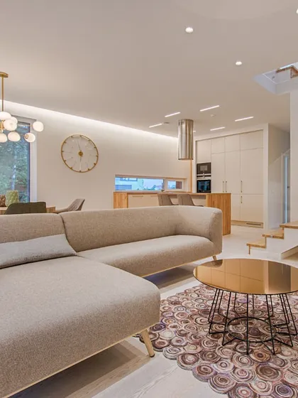 A bright and modern living room with an L-shaped sofa, an open kitchen in the background, and a stylish staircase. The neutral color palette and open layout make the space feel large and connected.
