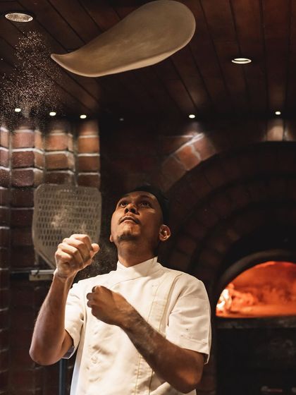 A chef in mid-toss, with a dusting of flour catching the light. This action shot captures the energy and craftsmanship that goes into every pizza we make.