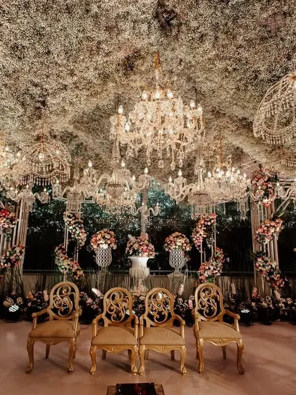 A view from within the mandap, looking out. The space is framed by elegant gold chairs for the couple and a ceiling dense with foliage and sparkling chandeliers.