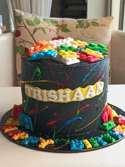A vibrant Lego-themed cake with a black base covered in colorful paint splatters and topped with edible building blocks.