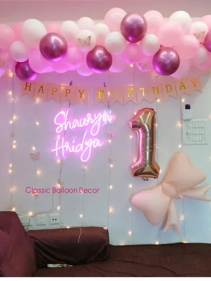 A chic first birthday wall decoration for a home party. It includes a balloon garland, a personalized neon sign, a '1' foil balloon, and a fairy light curtain.