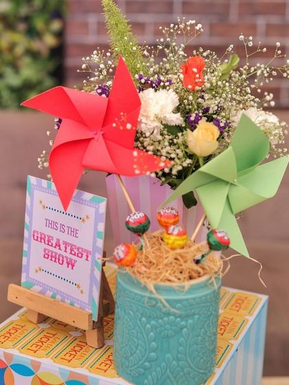 A charming carnival-themed centerpiece. This arrangement includes colorful pinwheels, lollipops, and fresh flowers, set against a mini "This is the Greatest Show" sign and a roll of tickets.