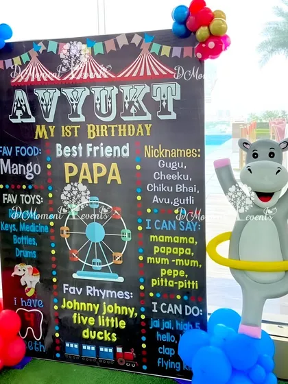 A personalized milestone chalkboard for Avyukt's first birthday carnival. The board lists all his favorites, from food to rhymes, next to a cute hula-hooping hippo balloon sculpture.