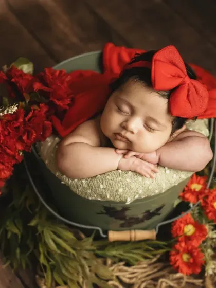 This angle captures the rich wooden background, contrasting beautifully with the baby's red accessories and the green bucket prop.