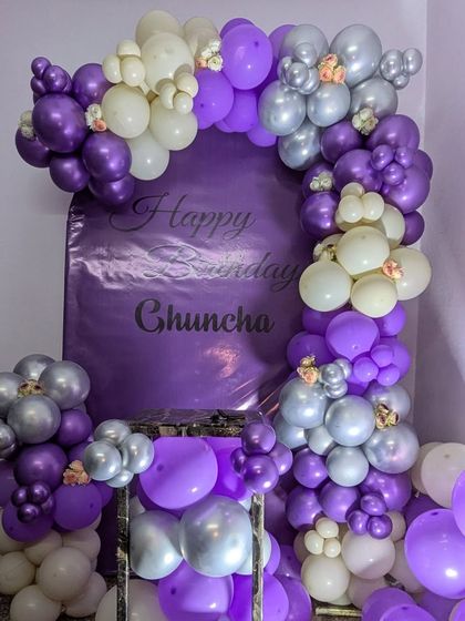 A custom balloon arch in purple, white, and silver with a personalized backdrop for a birthday celebration.