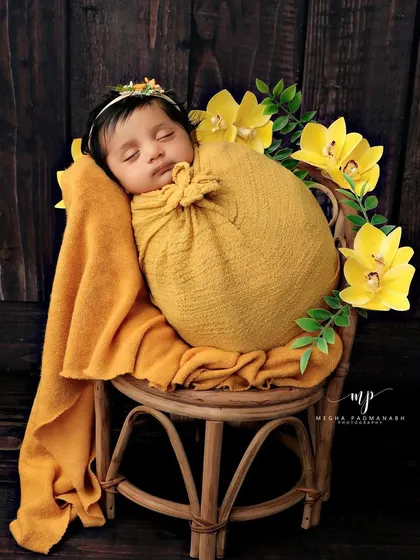 A baby in a yellow flower-style wrap, posed sitting up on a small rattan chair.