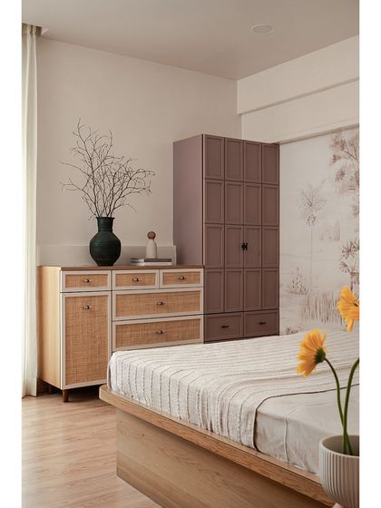 In this master bedroom, a cane-finished chest of drawers sits alongside a mauve-toned wardrobe, adding function and a soft splash of color. The walls are micro-topped for a seamless, textured finish.