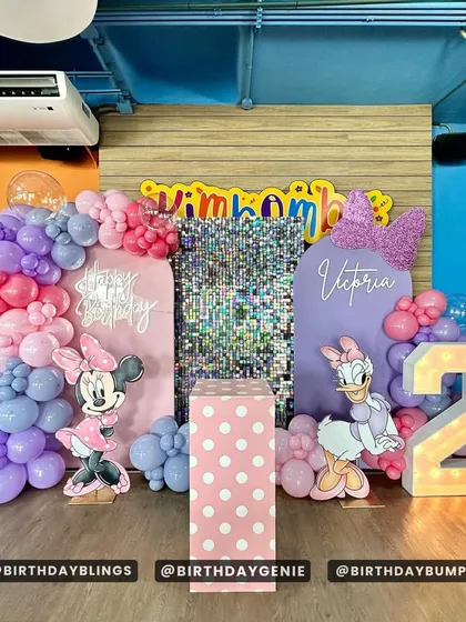 A Minnie and Daisy Duck party for a second birthday. The setup includes a purple and pink color scheme, a shimmer wall, and cutouts of both characters.