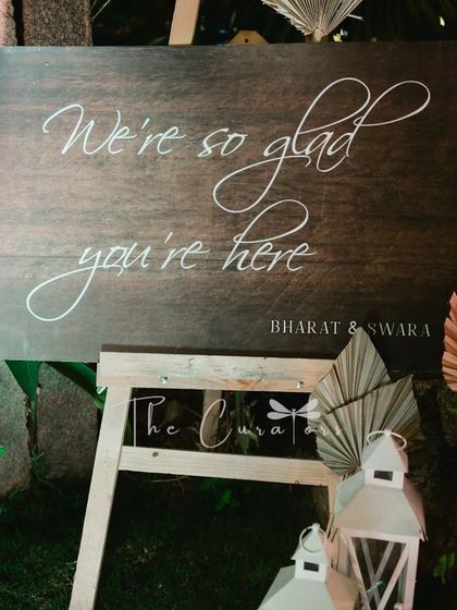"We're so glad you're here." A simple, heartfelt message on a rustic wooden sign, surrounded by pampas grass and soft pink roses, makes every guest feel welcome.