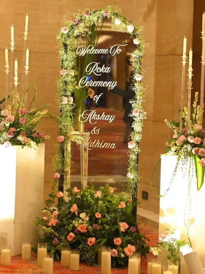 A personalized welcome for an intimate Roka. A floral-framed mirror with the couple's names is flanked by illuminated pedestals and candles, creating a warm and inviting entrance.