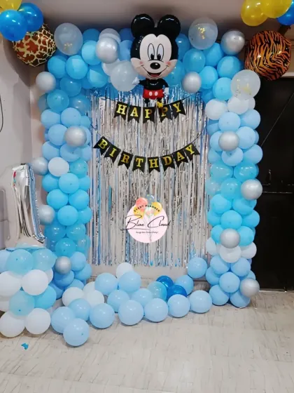 A Mickey Mouse first birthday setup with a blue and white balloon arch and a silver shimmer curtain.