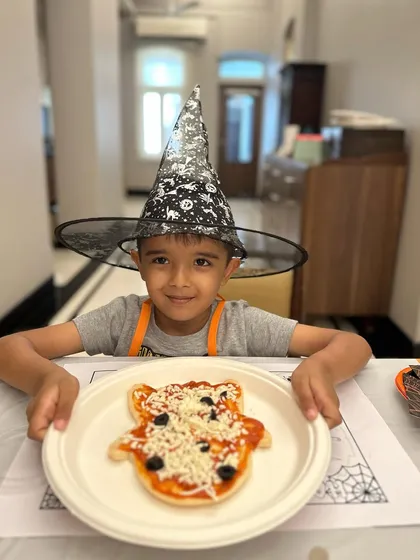 This little wizard is proudly presenting his ghost-shaped pizza, ready to be topped with cheese and olives.