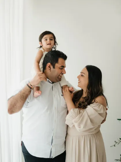 A happy family of three poses in the studio, with their little one perched on dad's shoulders. This is a classic pose for a simple lifestyle family photoshoot.