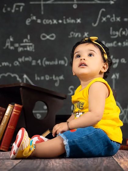 A baby girl looks up thoughtfully, as if pondering the complex equations on the chalkboard behind her. A cute and imaginative shot.