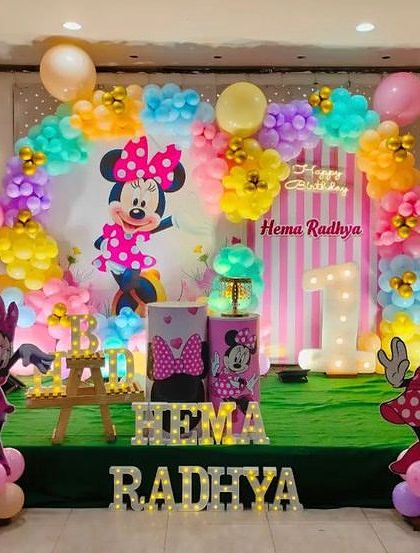 A cheerful Minnie Mouse first birthday stage. This setup features character cutouts, a colorful balloon arch, and light-up letters on a green turf stage.