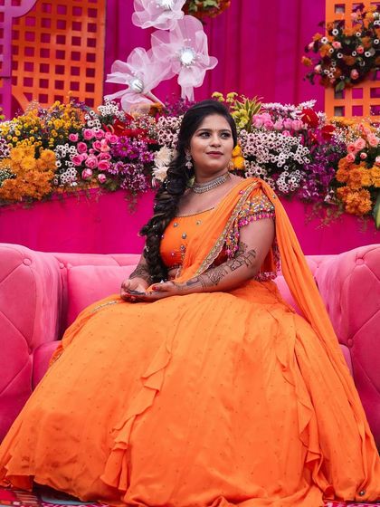 A beautiful bride in a vibrant orange lehenga, seated on a pink sofa, looking like a princess.