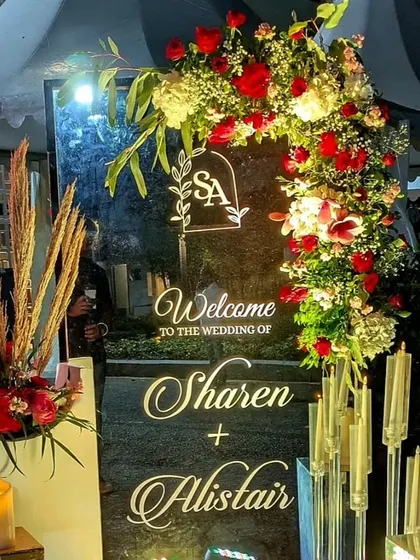 A personalized welcome is a beautiful touch. This custom acrylic sign for Sharen and Alistair features their monogram and is framed by a lush, asymmetrical arrangement of red and white roses, lit softly by candles for a romantic glow.