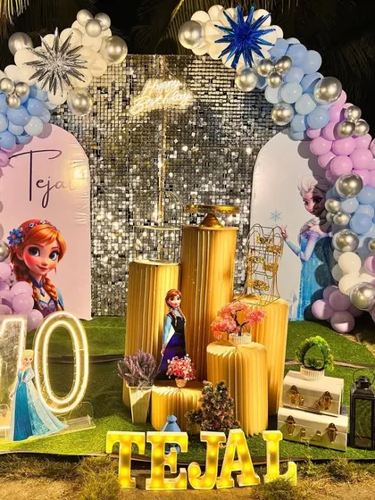 A magical outdoor 'Frozen' theme birthday party for a 10th birthday. The setup includes a silver shimmer wall, character cutouts of Anna and Elsa, and a balloon arch in pastel blues, purples, and whites, all brought together with custom light-up letters for the birthday girl's name.