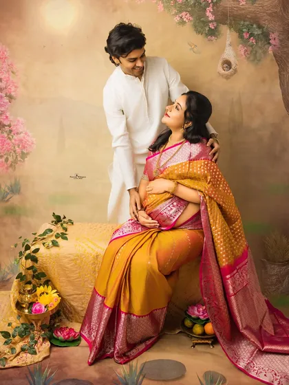 A serene and regal portrait of an expecting couple. The mother-to-be is seated gracefully while her partner stands by her side, creating a classic composition filled with love.