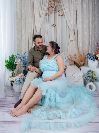 A romantic and dreamy maternity portrait. The couple is seated in a beautifully decorated boho-themed studio setup, holding hands and sharing a look of love and excitement.