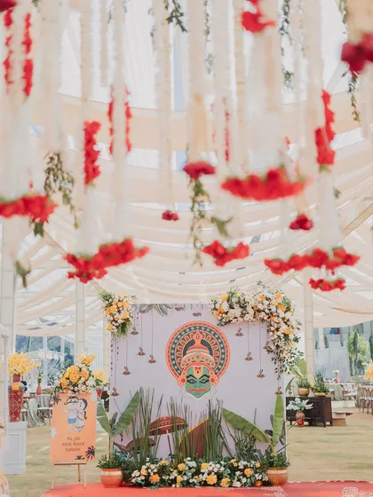 A culturally inspired backdrop featuring a Kathakali mask motif, perfect for a South Indian themed event. The hanging floral strings and banana leaf accents complete the traditional yet stylish look.