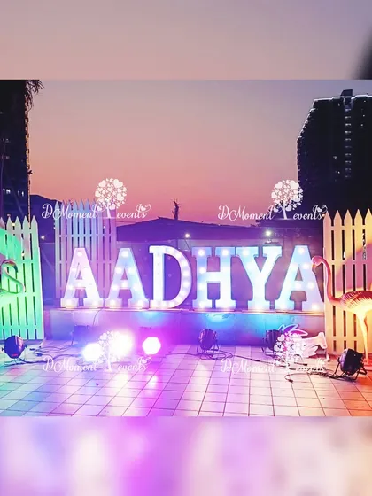 An outdoor setup for a Flamingo-themed 6th birthday party. Large, illuminated marquee letters spelling "AADHYA" are flanked by flamingo props and a white picket fence, creating a stylish photo op against the sunset.
