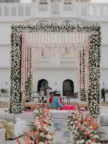 An elegant and understated mandap design, featuring a structure covered in dense white florals with delicate pink floral strings, set against the beautiful white architecture of the palace venue.