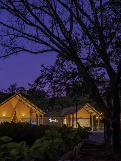 Teyla at dusk, with warm light spilling from its gabled structures. The building's silhouette against the evening sky creates a welcoming beacon for visitors, while the surrounding landscape is subtly lit.