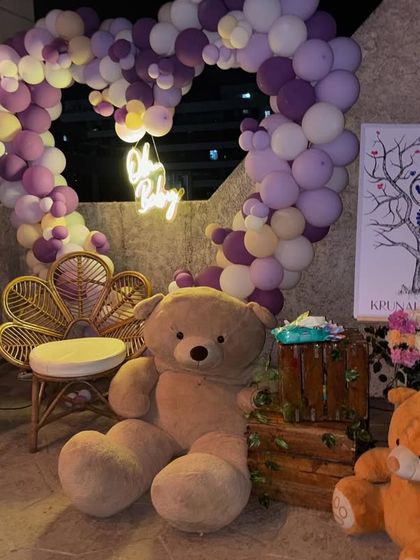 A cozy baby shower corner featuring a heart-shaped balloon arch in purple and white, a neon 'Oh Baby' sign, and large teddy bears.