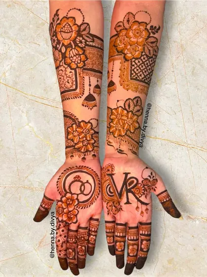 This personalized engagement mehendi features the couple's initials 'VR' and a symbol of intertwined rings. The design is complemented by floral patterns and hanging jhumka motifs.
