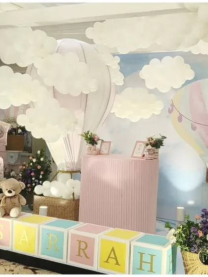 A whimsical hot air balloon themed party. The setup features a sky-blue backdrop, fluffy white clouds, and large name blocks spelling "SARRAH" in front of a pink dessert bar.