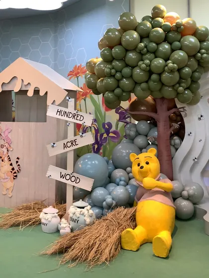 A full view of the Winnie the Pooh first birthday setup, featuring a large Pooh bear figure, a "Hundred Acre Wood" sign, and balloon trees in soft, natural colors.